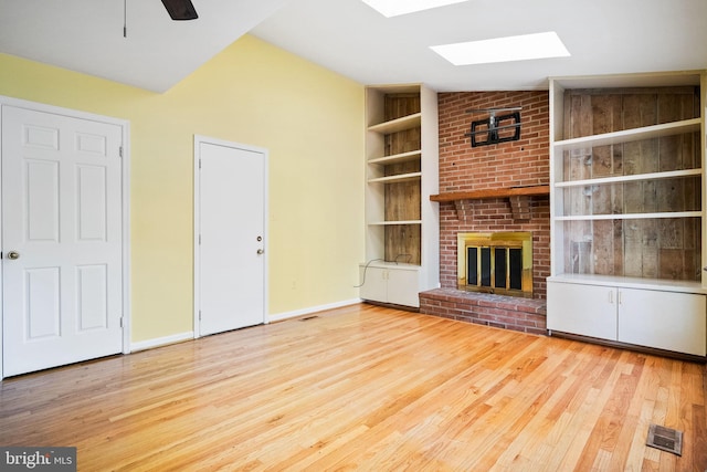 unfurnished living room featuring vaulted ceiling with skylight, ceiling fan, light wood-type flooring, and a brick fireplace