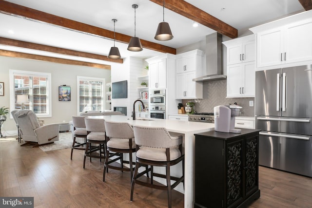 kitchen featuring beamed ceiling, a center island with sink, wall chimney range hood, white cabinetry, and appliances with stainless steel finishes