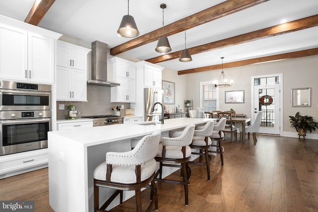 kitchen with a kitchen island with sink, dark hardwood / wood-style floors, pendant lighting, wall chimney range hood, and appliances with stainless steel finishes