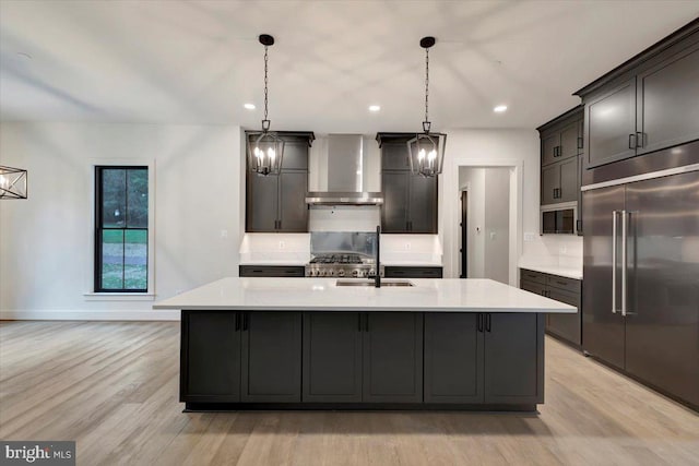 kitchen with wall chimney range hood, built in refrigerator, an island with sink, light hardwood / wood-style floors, and decorative light fixtures