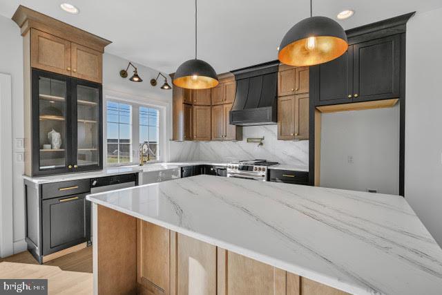 kitchen with light stone counters, custom exhaust hood, hanging light fixtures, and stainless steel stove