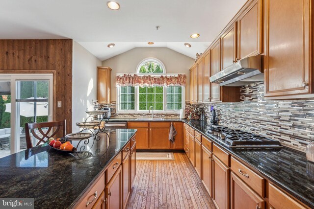 kitchen with stainless steel gas cooktop, backsplash, vaulted ceiling, dark stone countertops, and light hardwood / wood-style flooring