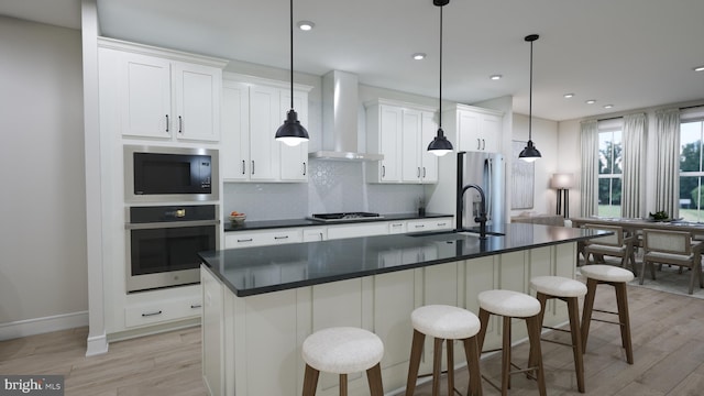 kitchen with an island with sink, white cabinets, wall chimney exhaust hood, sink, and appliances with stainless steel finishes