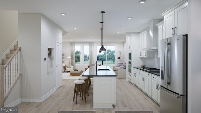 kitchen featuring stainless steel appliances, white cabinets, wall chimney range hood, hanging light fixtures, and a center island with sink