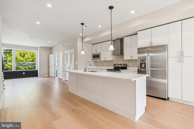 kitchen with wall chimney range hood, white cabinets, an island with sink, appliances with stainless steel finishes, and sink