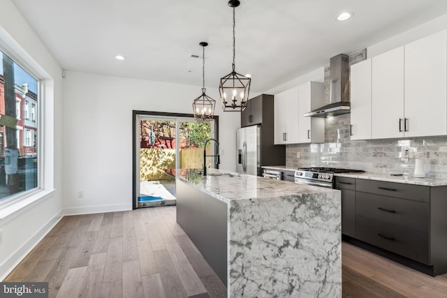 kitchen featuring decorative light fixtures, an island with sink, wood-type flooring, white cabinets, and wall chimney range hood