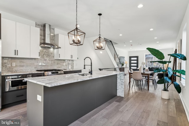 kitchen featuring stainless steel appliances, white cabinetry, hanging light fixtures, light hardwood / wood-style floors, and wall chimney exhaust hood