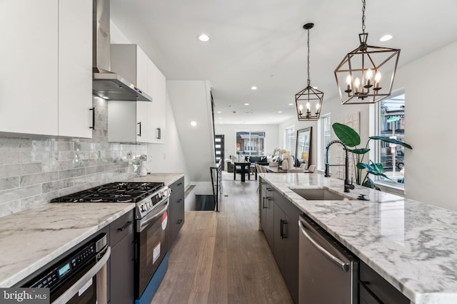 kitchen featuring wall chimney range hood, appliances with stainless steel finishes, hanging light fixtures, sink, and white cabinets