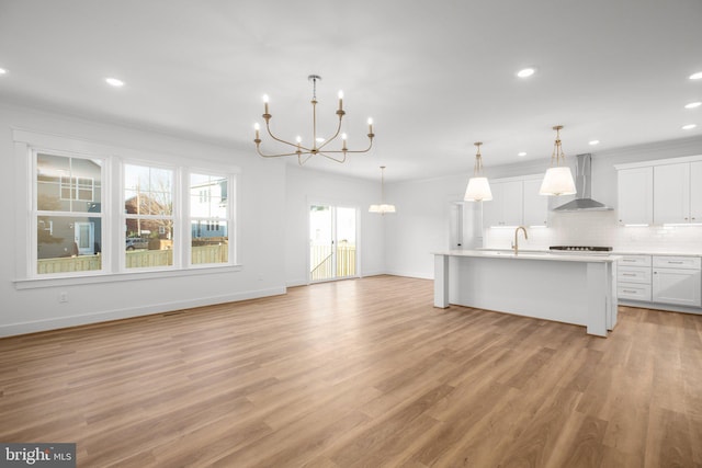 kitchen with white cabinetry, wall chimney range hood, light hardwood / wood-style floors, decorative light fixtures, and a kitchen island with sink