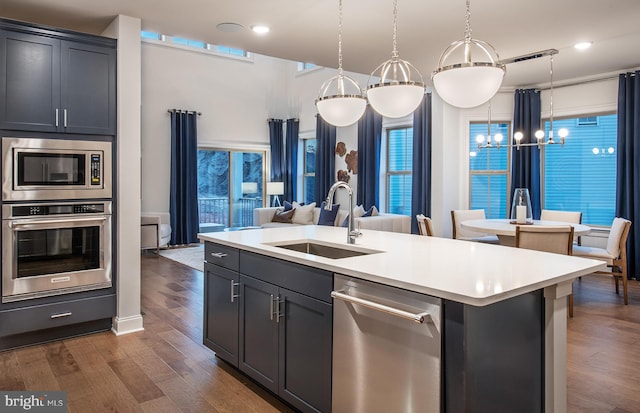 kitchen with dark wood-type flooring, a center island with sink, sink, hanging light fixtures, and appliances with stainless steel finishes