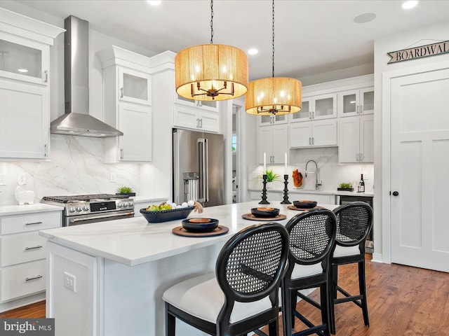 kitchen featuring an island with sink, hardwood / wood-style floors, wall chimney range hood, white cabinetry, and appliances with stainless steel finishes