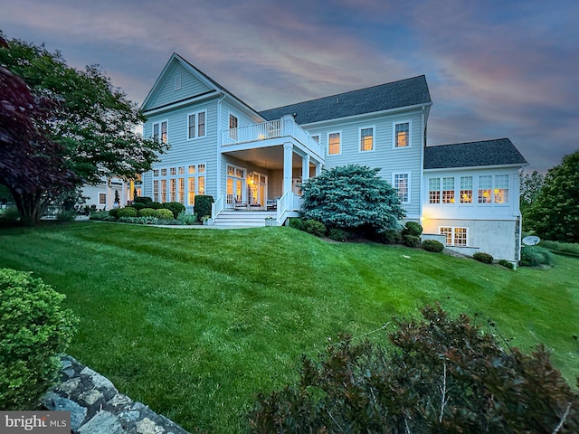 back of house at dusk featuring a lawn and a balcony