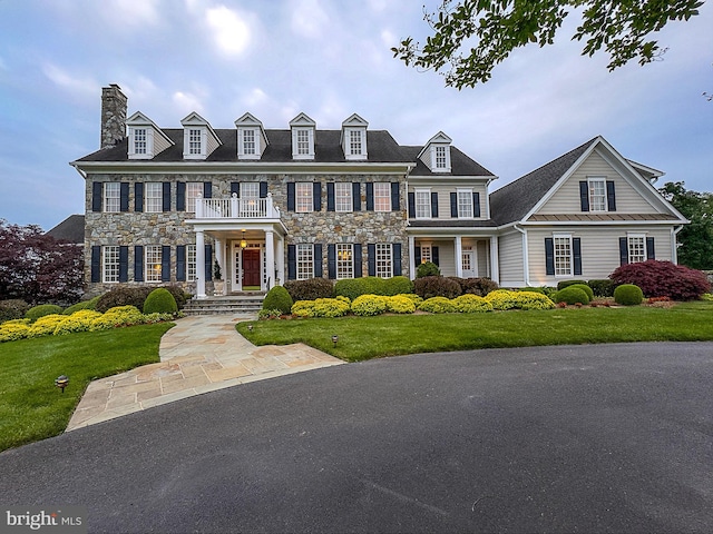 colonial-style house with a chimney, a front lawn, stone siding, and a balcony