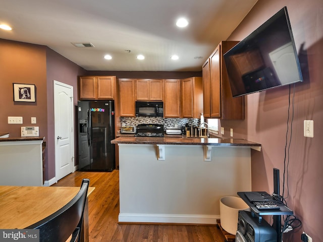 kitchen with black appliances, hardwood / wood-style flooring, kitchen peninsula, dark stone counters, and a breakfast bar area