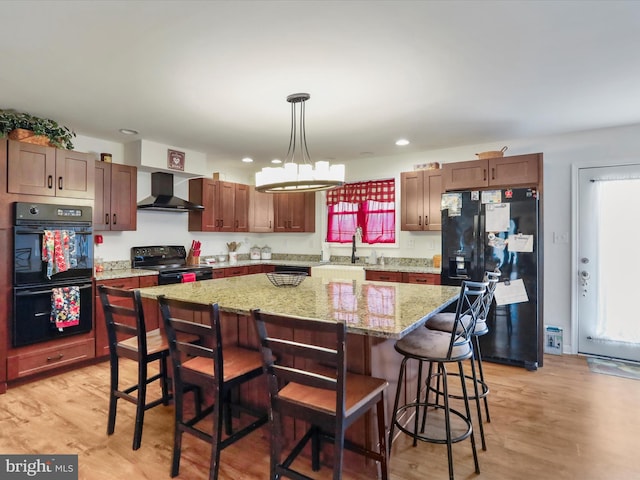 kitchen featuring black appliances, light wood-type flooring, a kitchen island, wall chimney exhaust hood, and decorative light fixtures