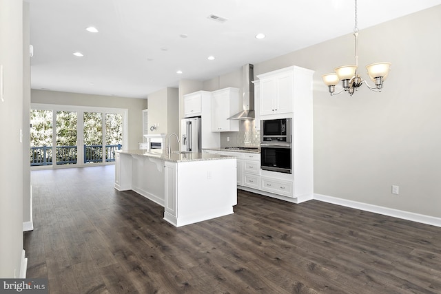 kitchen featuring light stone countertops, wall chimney range hood, white cabinetry, appliances with stainless steel finishes, and a center island with sink