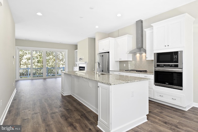 kitchen with wall chimney exhaust hood, dark wood-type flooring, a center island with sink, white cabinets, and appliances with stainless steel finishes