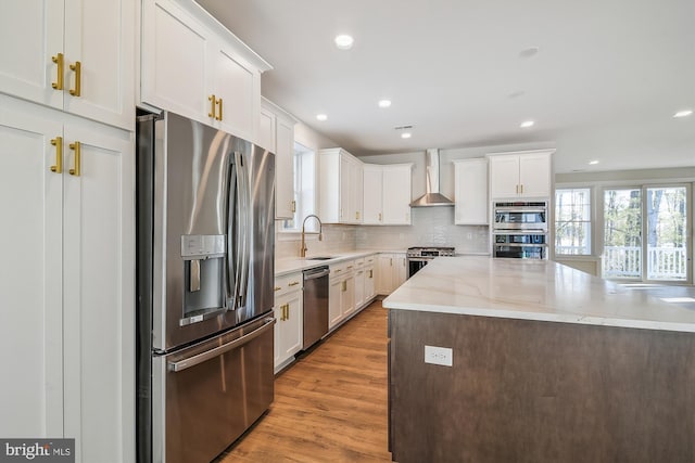 kitchen with stainless steel appliances, decorative backsplash, a sink, light wood-type flooring, and wall chimney exhaust hood