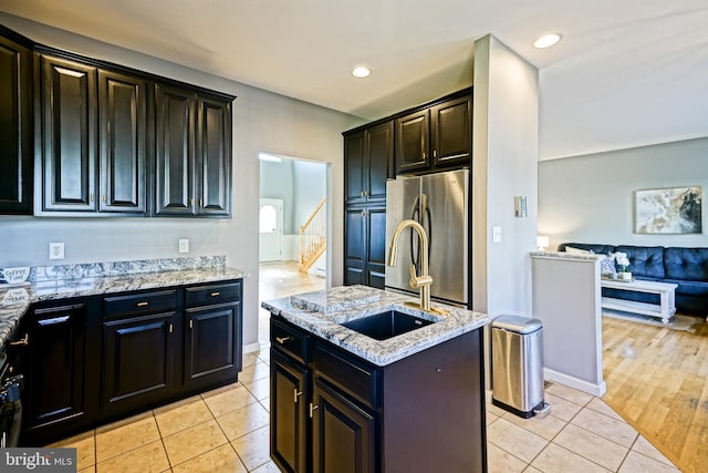 kitchen with light tile patterned floors, a sink, freestanding refrigerator, an island with sink, and light stone countertops
