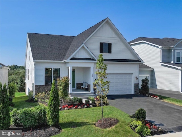 view of front of home with a front lawn, stone siding, covered porch, a shingled roof, and aphalt driveway