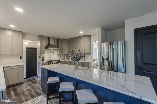 kitchen featuring a breakfast bar area, appliances with stainless steel finishes, dark wood-type flooring, wall chimney exhaust hood, and light stone counters