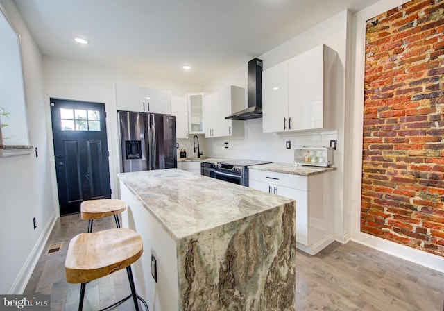 kitchen featuring wall chimney exhaust hood, light stone countertops, a kitchen bar, white cabinetry, and appliances with stainless steel finishes