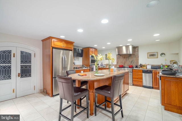 kitchen featuring appliances with stainless steel finishes, a kitchen island, wall chimney exhaust hood, light stone counters, and a breakfast bar area