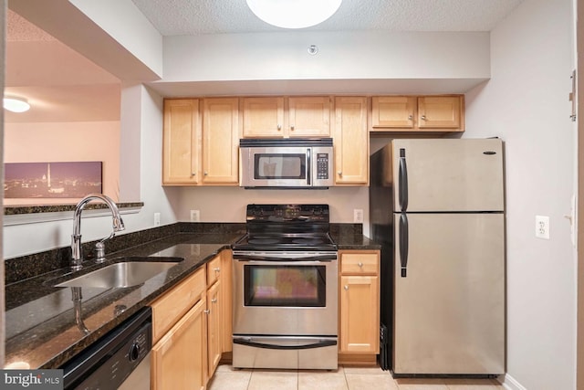 kitchen featuring appliances with stainless steel finishes, light brown cabinetry, dark stone countertops, light tile patterned flooring, and sink