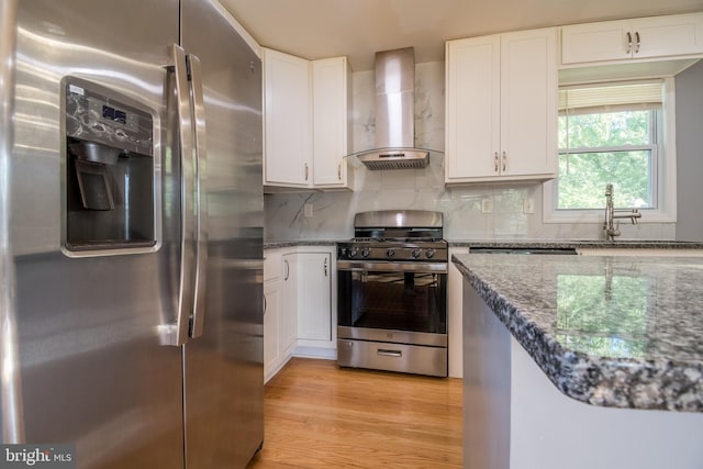 kitchen with wall chimney range hood, appliances with stainless steel finishes, white cabinetry, light hardwood / wood-style floors, and dark stone countertops