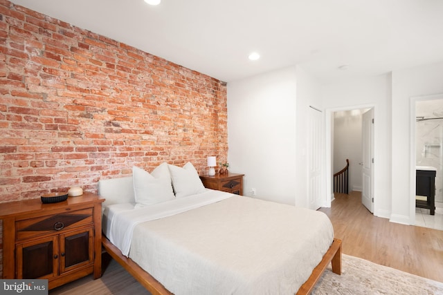 bedroom with light wood finished floors, baseboards, recessed lighting, brick wall, and connected bathroom