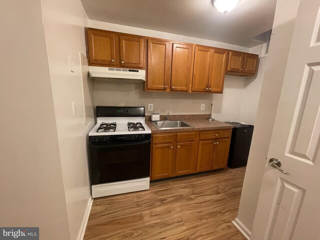 kitchen with under cabinet range hood, brown cabinetry, a sink, gas range oven, and light wood-style flooring