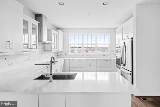 kitchen with wall chimney range hood, kitchen peninsula, sink, white cabinets, and appliances with stainless steel finishes