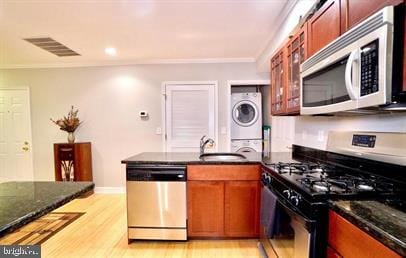 kitchen featuring sink, light hardwood / wood-style floors, stainless steel appliances, dark stone counters, and stacked washer and clothes dryer