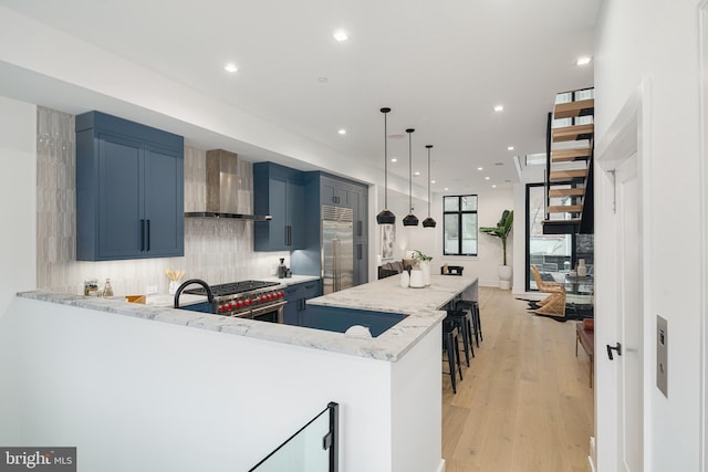 kitchen with wall chimney range hood, high end appliances, light stone countertops, light wood-type flooring, and blue cabinets