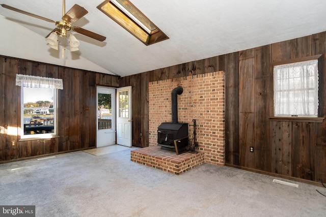 unfurnished living room featuring ceiling fan, wood walls, vaulted ceiling with skylight, and a wood stove