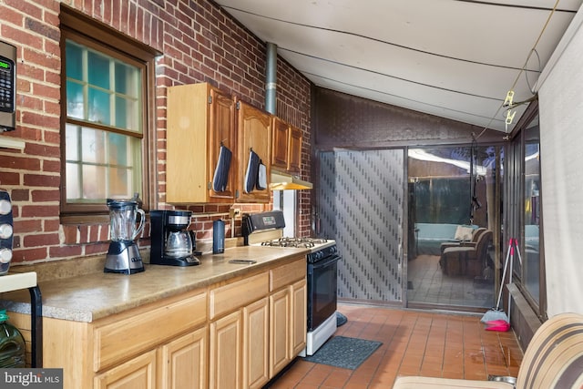 kitchen with brick wall, light brown cabinetry, gas range gas stove, and vaulted ceiling