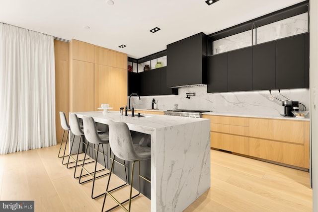 kitchen with sink, light stone counters, an island with sink, light brown cabinetry, and wall chimney range hood