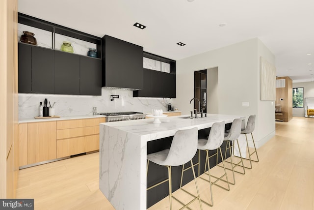 kitchen with a center island with sink, light stone countertops, light brown cabinetry, sink, and wall chimney range hood