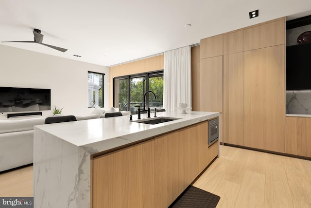 kitchen with sink, light brown cabinets, ceiling fan, an island with sink, and light stone countertops