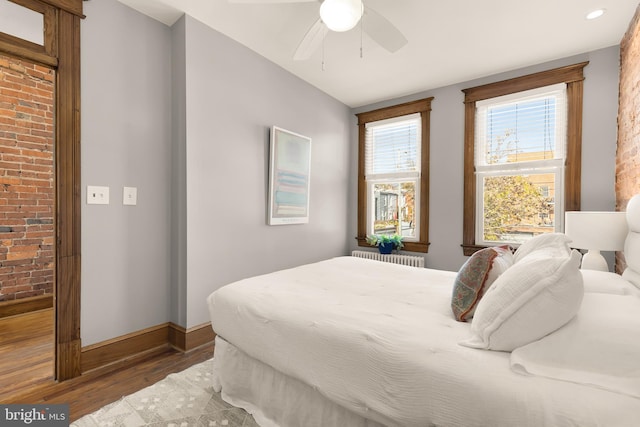 bedroom with hardwood / wood-style flooring, ceiling fan, radiator heating unit, and brick wall