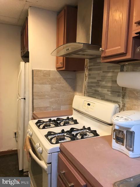 kitchen featuring brown cabinetry, decorative backsplash, wall chimney exhaust hood, and white appliances