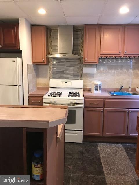 kitchen with a sink, white appliances, light countertops, wall chimney exhaust hood, and a paneled ceiling