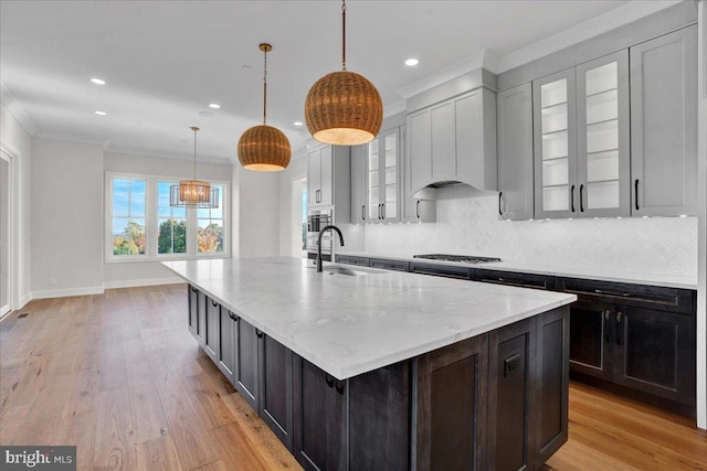 kitchen featuring sink, light wood-type flooring, stainless steel gas cooktop, hanging light fixtures, and a center island with sink