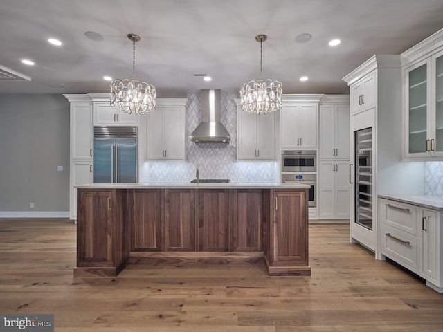 kitchen with wall chimney range hood, hanging light fixtures, appliances with stainless steel finishes, and a kitchen island with sink