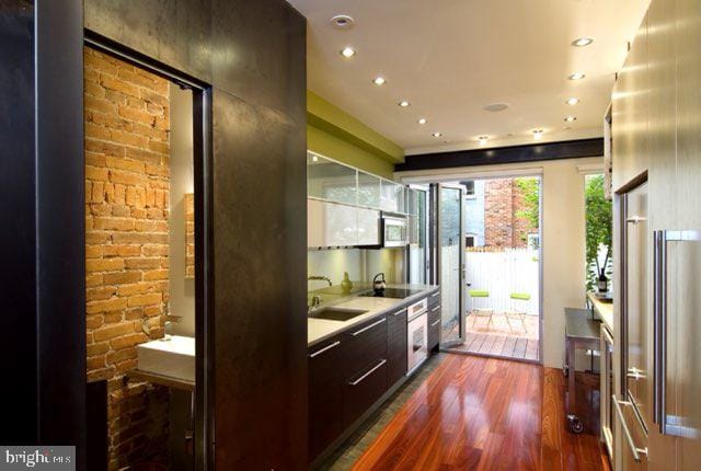 kitchen with dark hardwood / wood-style flooring, brick wall, cooktop, dark brown cabinetry, and sink