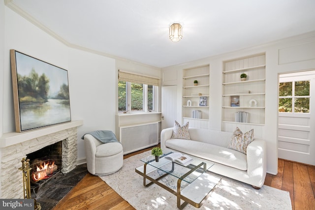 living room with a stone fireplace, hardwood / wood-style flooring, ornamental molding, built in features, and radiator heating unit