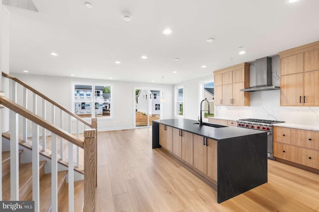 kitchen with high end stove, a kitchen island with sink, wall chimney range hood, sink, and light hardwood / wood-style floors