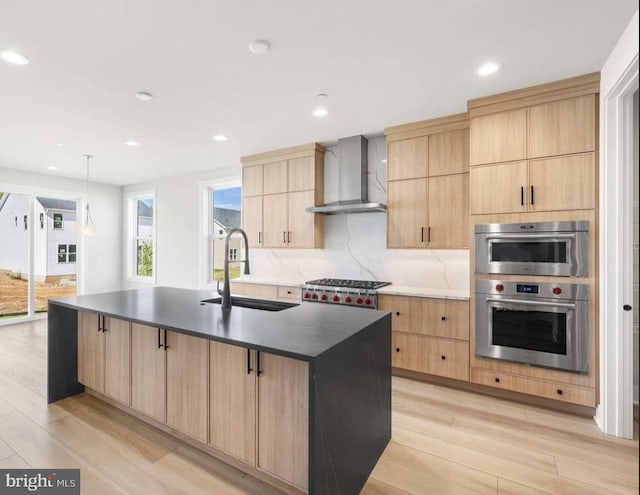 kitchen with wall chimney range hood, sink, stainless steel appliances, light hardwood / wood-style flooring, and a kitchen island with sink