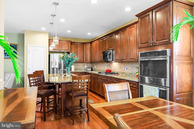 kitchen with wood-type flooring, appliances with stainless steel finishes, light stone countertops, hanging light fixtures, and an island with sink