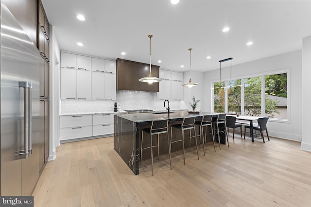 kitchen with stainless steel built in fridge, white cabinets, hanging light fixtures, a center island with sink, and light hardwood / wood-style flooring